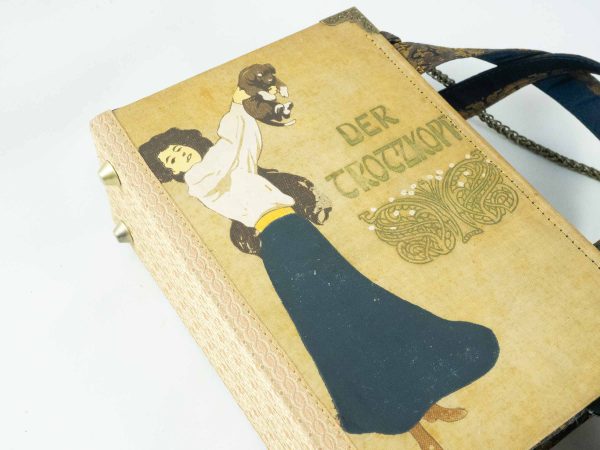 Tasche aus einem alten Buch "Der Trotzkopf" in beige. Am Cover ist eine Frau, die ein Hündchen in die Höhe hält, zu sehen. Buch kombiniert mit einer dunkelblau/goldenen Krawatte und goldenem Stoff.