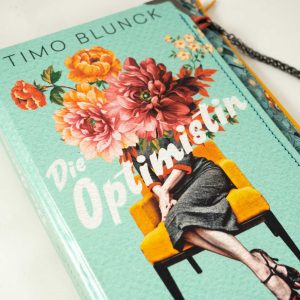 Tasche/Clutch aus einem Buch "Die Optimistin" mit einer Frau am Cover, Sitzens auf einem Stuhl. Das Gesicht ist mit Blumen verdeckt. Buch ist in hellblau/helltürkies gehalten. Das Buch ist mit ebenso farblich gehaltenen Krawatte kombiniert.