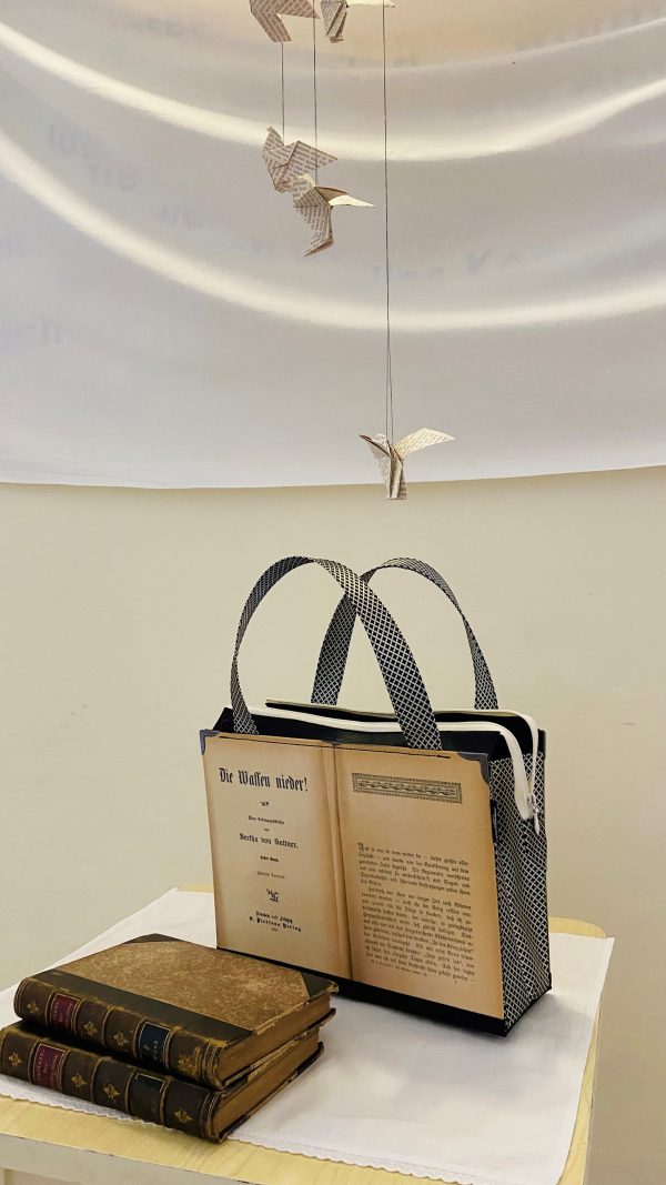 Tasche aus Buchseiten von Bertha von Suttners Buch "Die Waffen nieder" mit darüber schwebenden Friedenstauben aus Buchseiten gefaltet