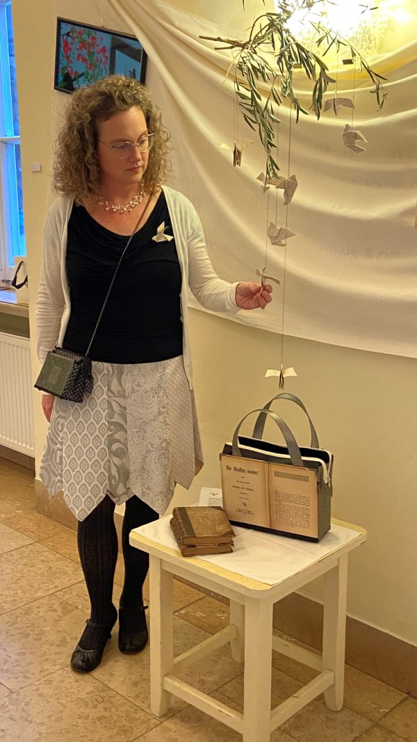 Bernadette Hartl mit der Tasche aus Buchseiten von Bertha von Suttners Buch "Die Waffen nieder" mit darüber schwebenden Friedenstauben aus Buchseiten gefaltet